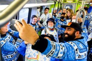 Man takes selfie of group wearing San Francisco BART holiday sweaters while riding BART.