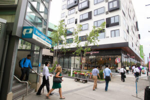 People exit Harrison PATH Station onto sidewalk amid mixed use development.