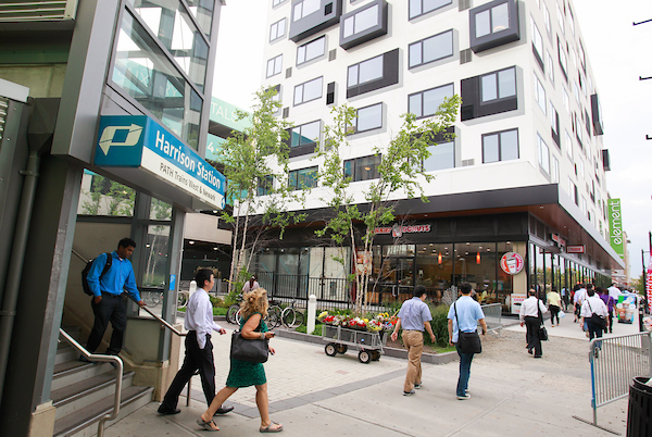 People exit Harrison PATH Station onto sidewalk amid mixed use development.