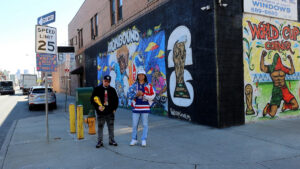 Two men, one hold replica World Cup trophy, stand in front of World Cup mural they created in Newark, New Jersey.