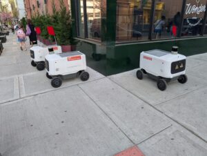 Avride delivery robots parked along sidewalk on street corner.