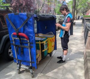 Delivery worker pauses on sidewalk with cart full of packages ordered via e-commerce.