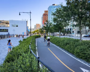 People ride along bike path between road and pedestrian walkway along river near Chelsea Piers.